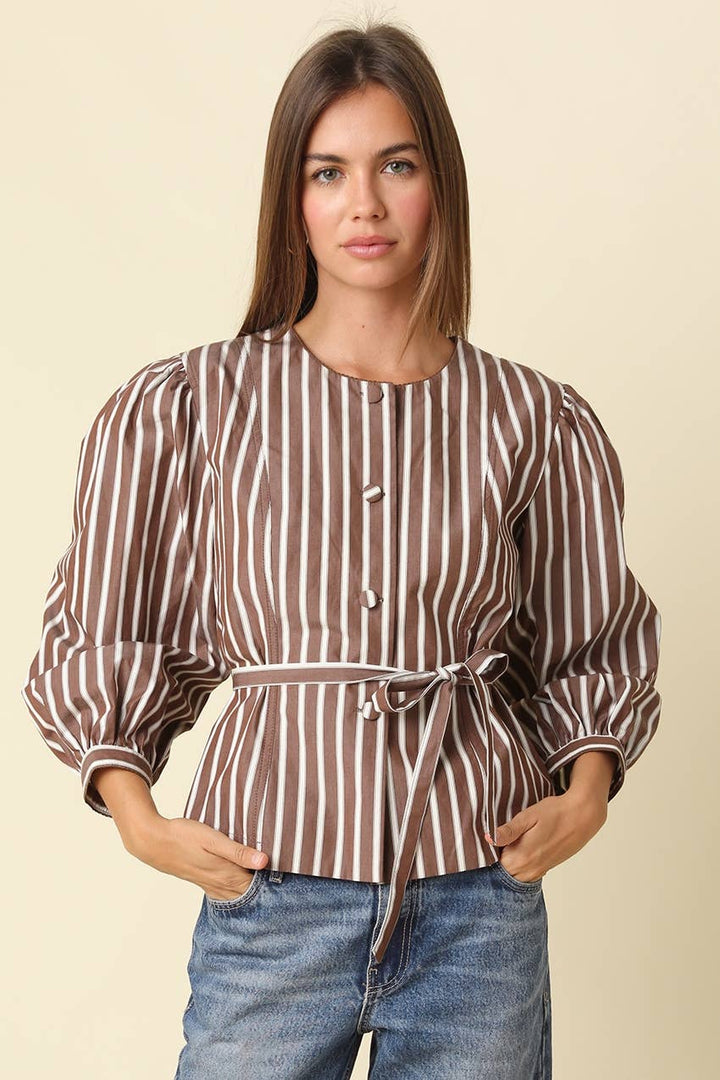 A woman modeling a brown and white vertically striped peplum blouse with puff sleeves and a tie waist, paired with blue jeans, against a solid beige background.