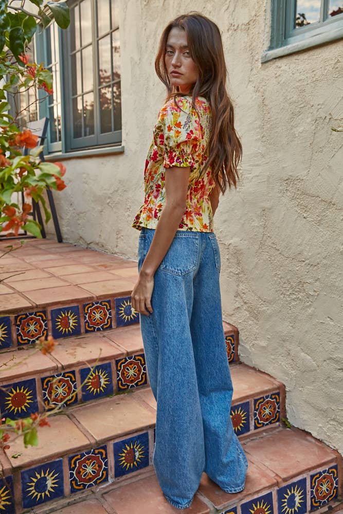 Woman in a multicolor floral puff sleeve top and blue jeans standing on a tiled staircase with a building in the background.