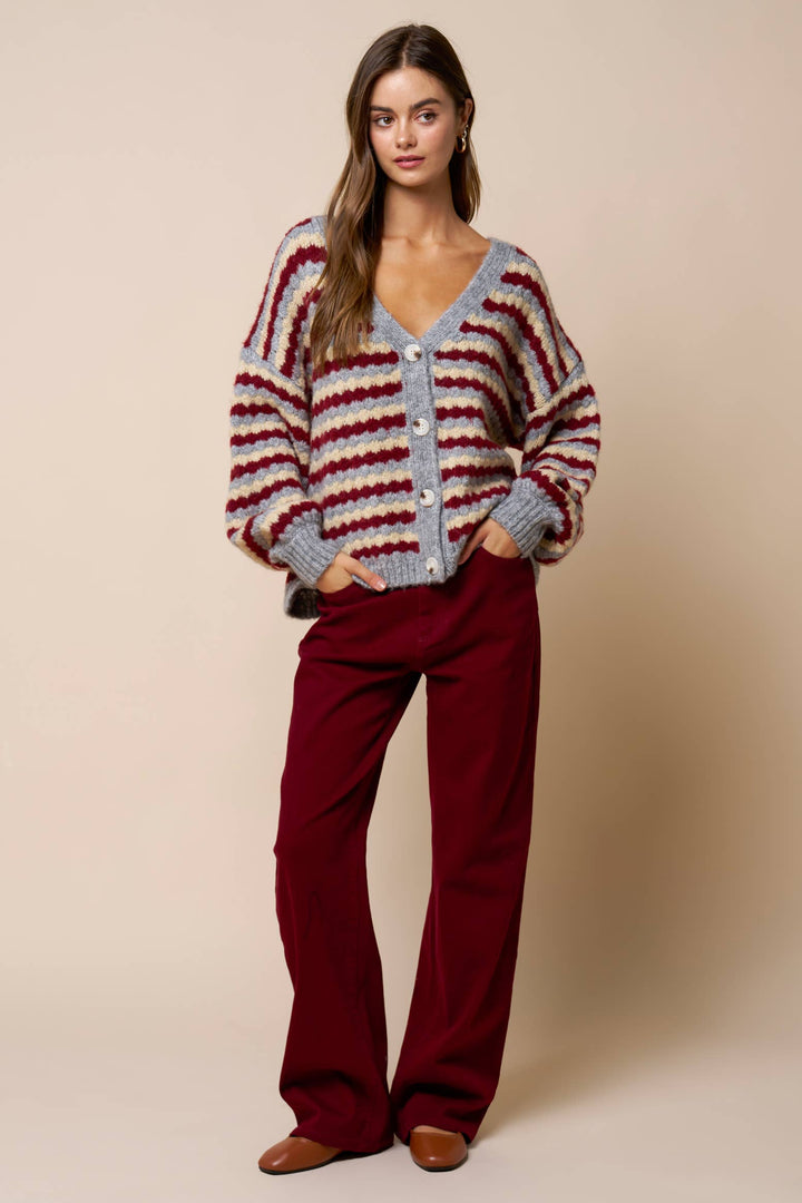 Woman wearing a striped gray and mulberry colored cardigan and red pants against a beige background
