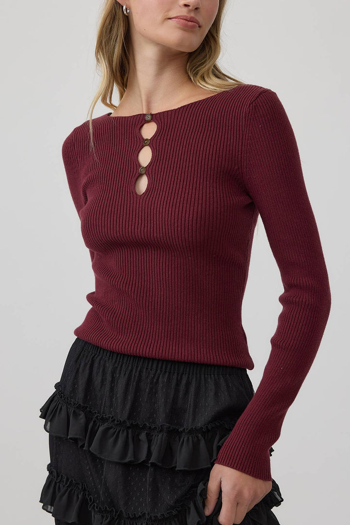 A woman wearing a ribbed long-sleeve knit top in deep burgundy with three decorative cutout details on the chest, paired with a black ruffled skirt, against a light gray studio background.