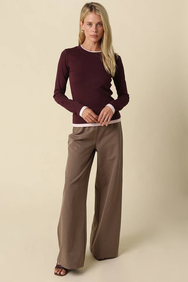 A blonde woman modeling a long-sleeve crewneck long sleeve tee in deep burgundy with light pink ribbed trim, paired with wide-leg taupe pants, against a solid beige studio background.