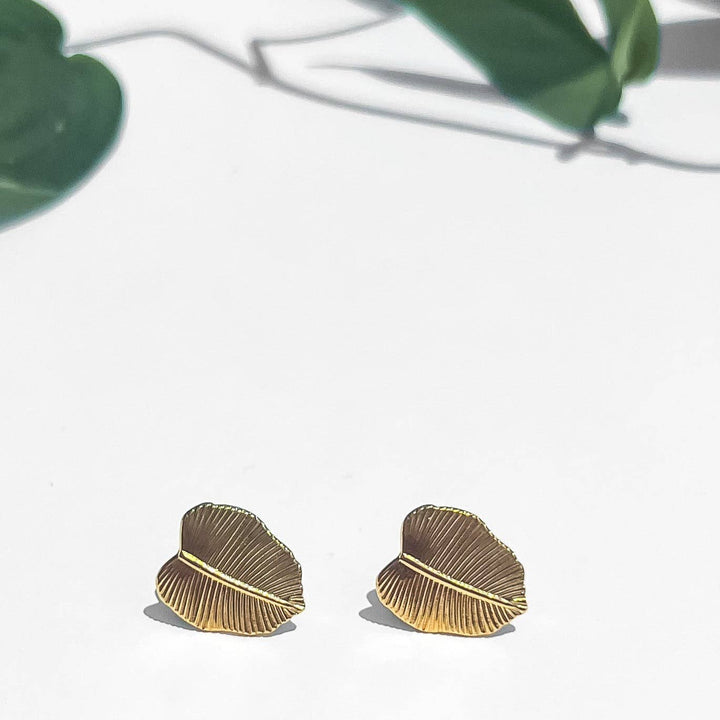 Gold leaf-shaped earrings on a white surface with green leaves in the background
