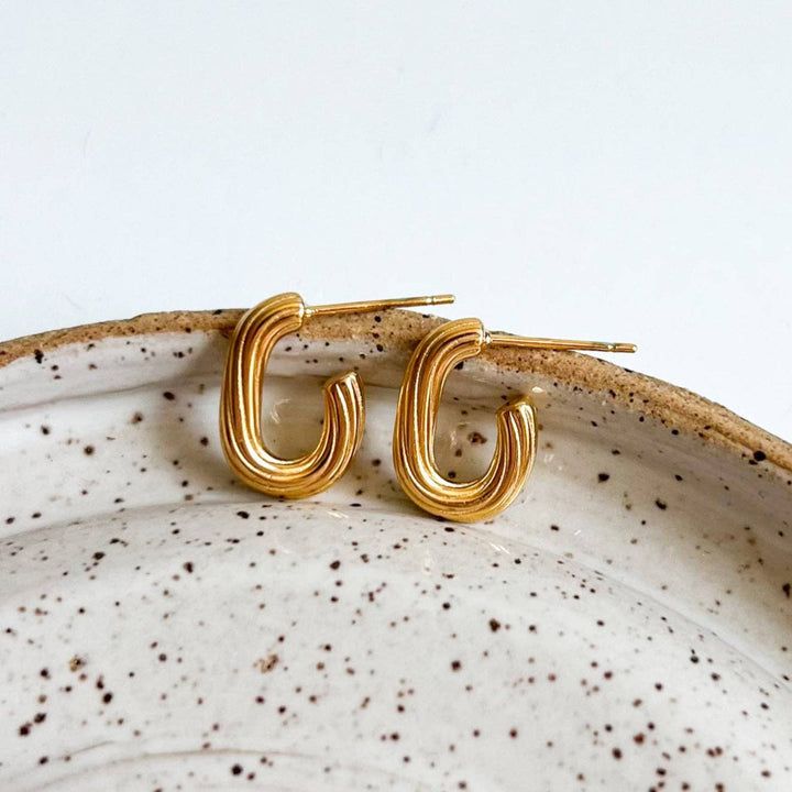 A pair of gold-toned, curved hoop earrings resting on the rim of a speckled ceramic dish against a clean white background.