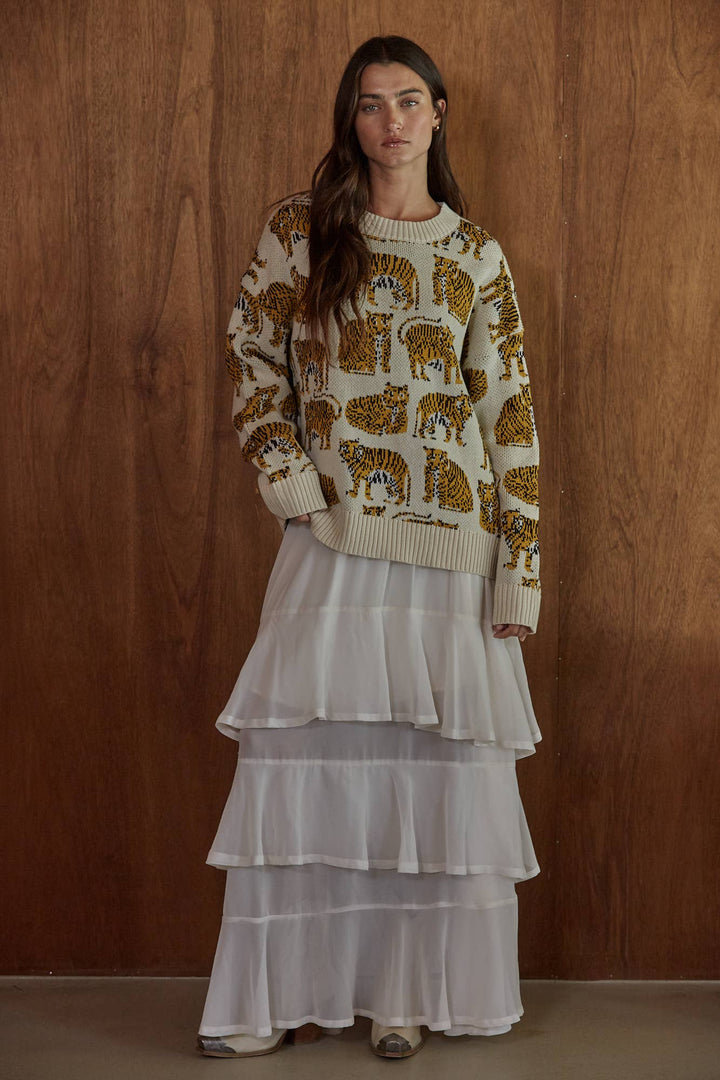 A woman stands against a warm wood-paneled wall wearing a cream knit sweater with a repeating tiger print and a long white tiered skirt.