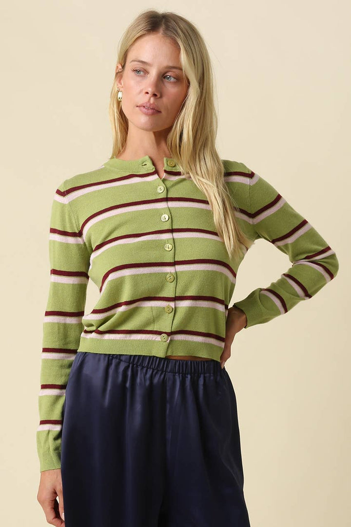 A woman modeling a green striped button-up cardigan with burgundy and cream horizontal stripes, paired with dark navy pants, against a solid beige background.