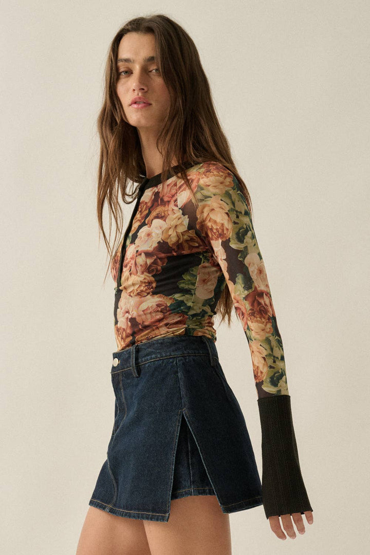A woman modeling a long-sleeve floral mesh top with black cuffs and a black collar, paired with a dark denim skirt, against a plain beige background.