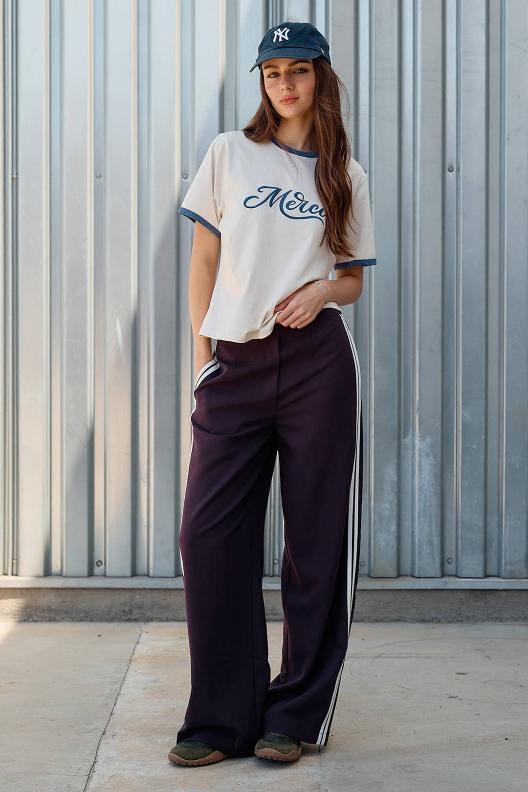 A woman stands outdoors against a corrugated metal wall wearing a cream ringer T-shirt with navy trim and script lettering, dark wide-leg track pants with white side stripes, and a navy baseball cap.