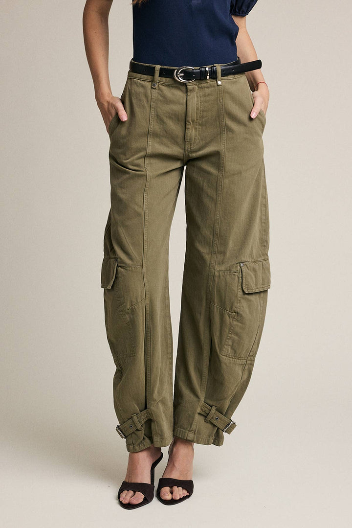 A woman wearing olive green wide-leg cargo pants with side flap pockets and bungee cuffs, styled with a black belt, navy top, and black sandals against a plain beige studio background.