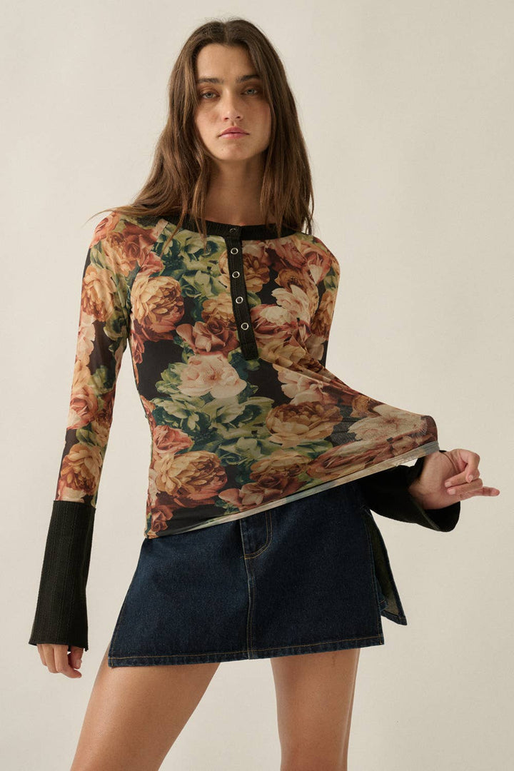 A woman modeling a long-sleeve floral top with black bell sleeves and a buttoned neckline, paired with a dark denim mini skirt, against a plain light beige studio background.