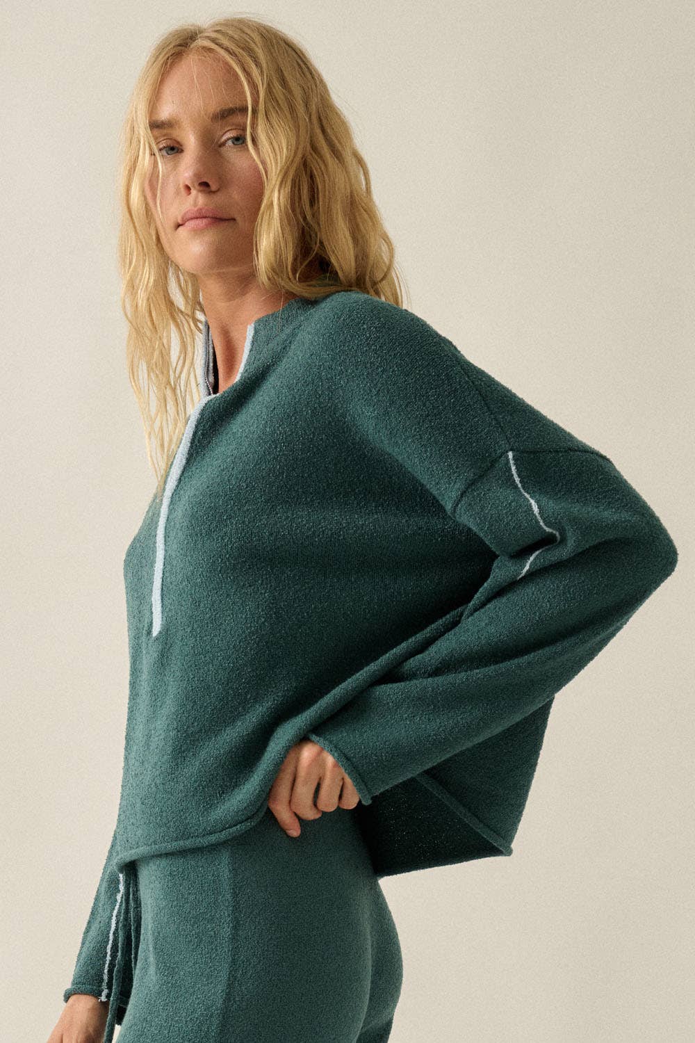 A woman wearing a dark teal green knit half-zip sweater and matching knit pants, photographed against a plain light beige studio background.