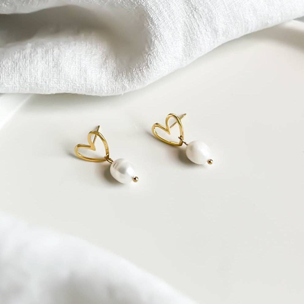 A pair of gold heart-shaped hoop earrings with dangling white pearl beads, displayed on a clean white surface with soft white fabric in the background.