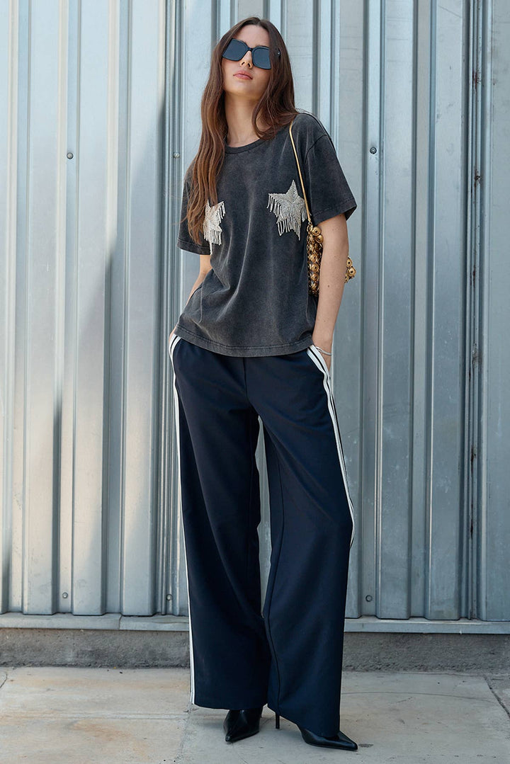 Woman wearing a dark gray t-shirt with star patterns and wide-leg navy pants against a metallic wall.