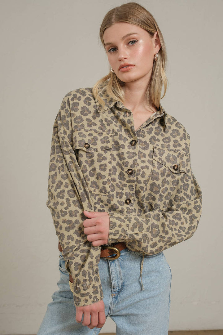 A woman wearing a beige and brown leopard-print button-up shirt with light blue jeans, standing against a neutral light gray wall.