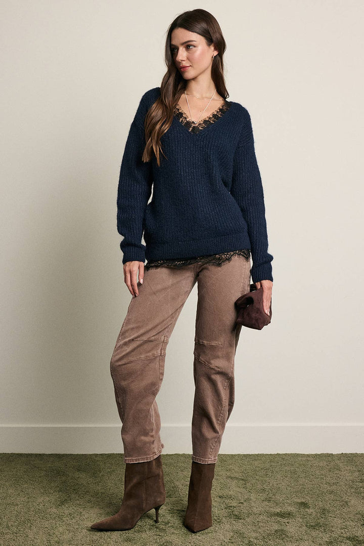 A woman modeling a navy blue ribbed knit sweater with a lace-trimmed V-neck, paired with brown pants and brown heeled ankle boots, standing against a plain beige wall.