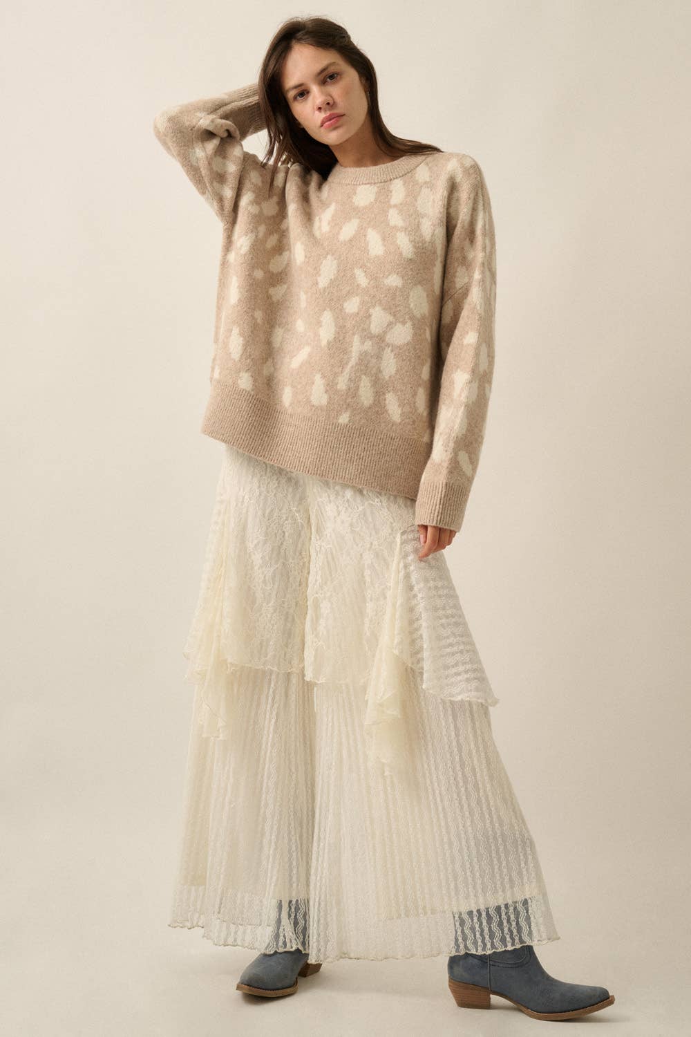 A woman modeling a beige knit sweater with a white abstract pattern, paired with a long cream pleated skirt and blue-gray ankle boots, against a plain beige studio background.