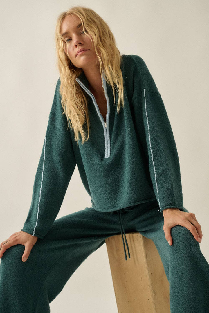A woman wearing a dark teal green knit lounge set with a mock neck and half-zip pullover and matching sweater pants, sitting on a wooden block against a plain light beige background.