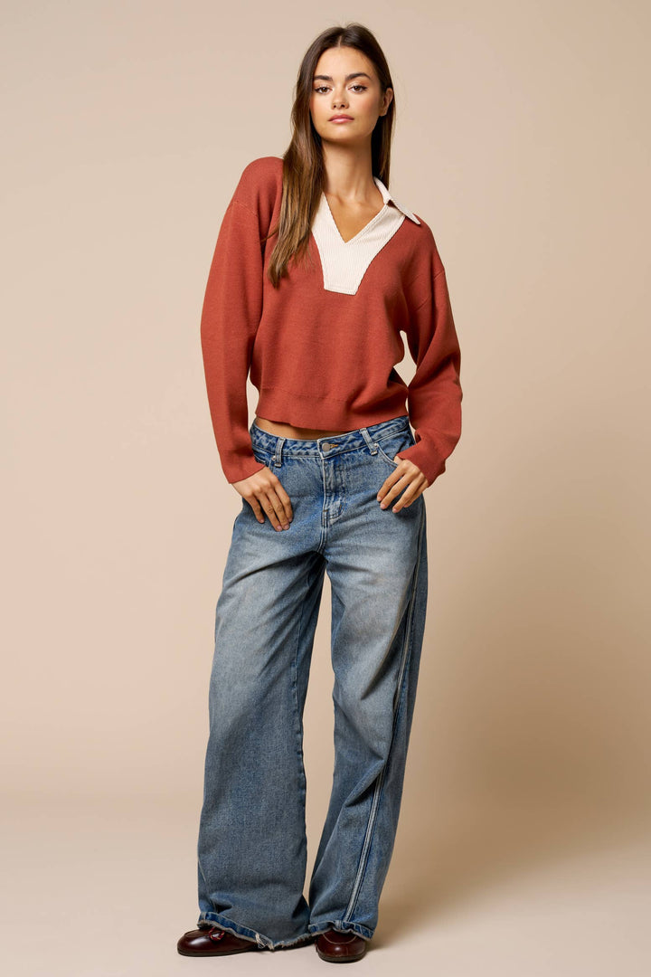 A woman modeling a rust-colored v-neck light weight knit sweater with a contrasting white collar, paired with wide-leg blue jeans and dark brown shoes, against a solid beige studio background.