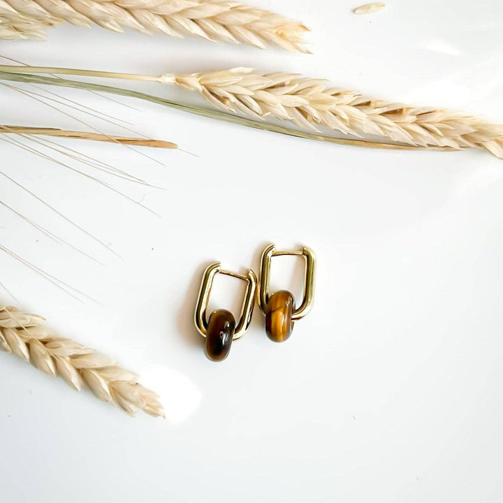 A pair of gold-tone hoop earrings with smooth, polished brown and golden tiger's eye stones, styled on a white surface with dried wheat stalks.