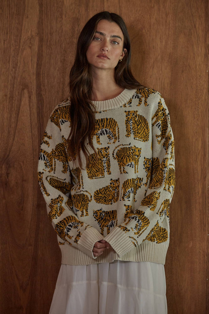 A woman wearing a cream-colored knit sweater with an all-over tiger print in brown, black, and orange, posed against a dark wood-paneled wall.
