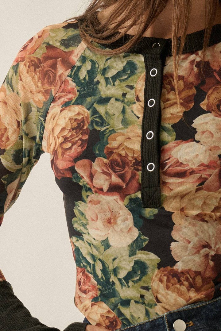 Close-up of a woman wearing a long-sleeve floral top with a black Henley-style placket featuring three silver eyelets, paired with dark denim bottoms.