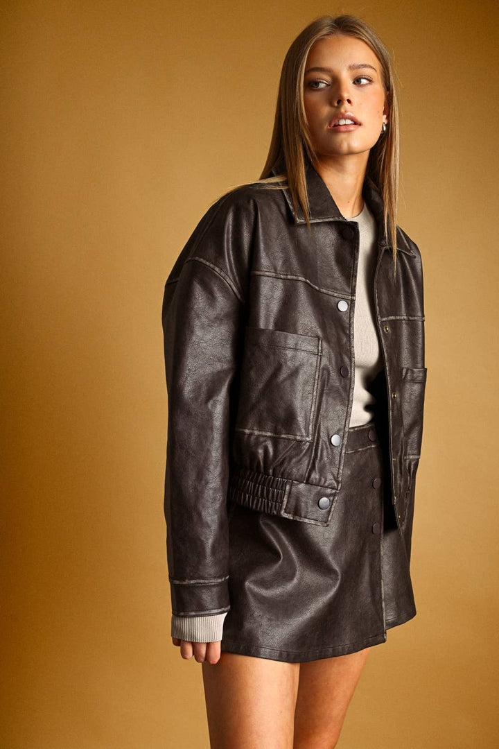 A woman modeling a dark brown faux-leather jacket and matching mini skirt set against a warm, solid mustard-yellow studio background.