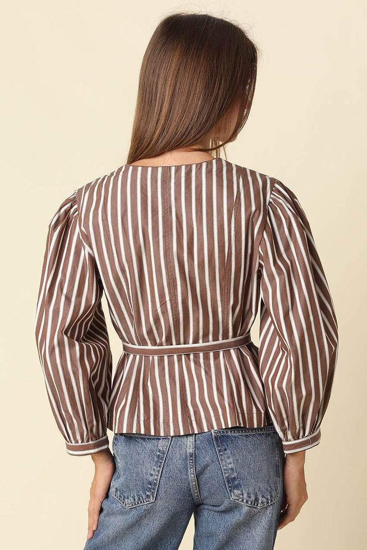A woman viewed from the back wearing a brown and white vertically striped peplum blouse with puffed long sleeves and a matching belt, paired with blue jeans against a solid light beige background.
