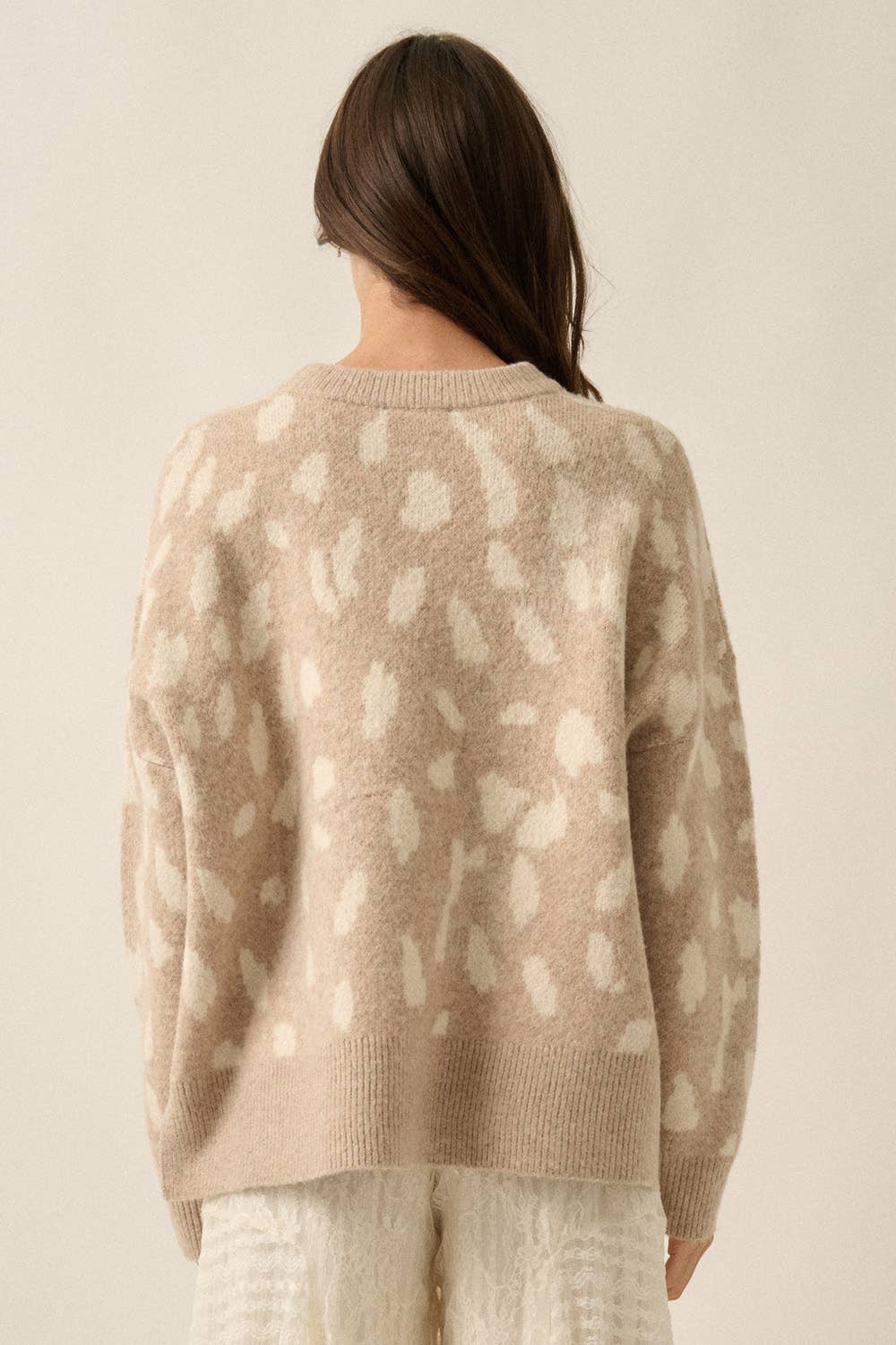 A woman viewed from the back wearing a cream and beige animal-print knit sweater and textured cream pants against a plain beige studio background.