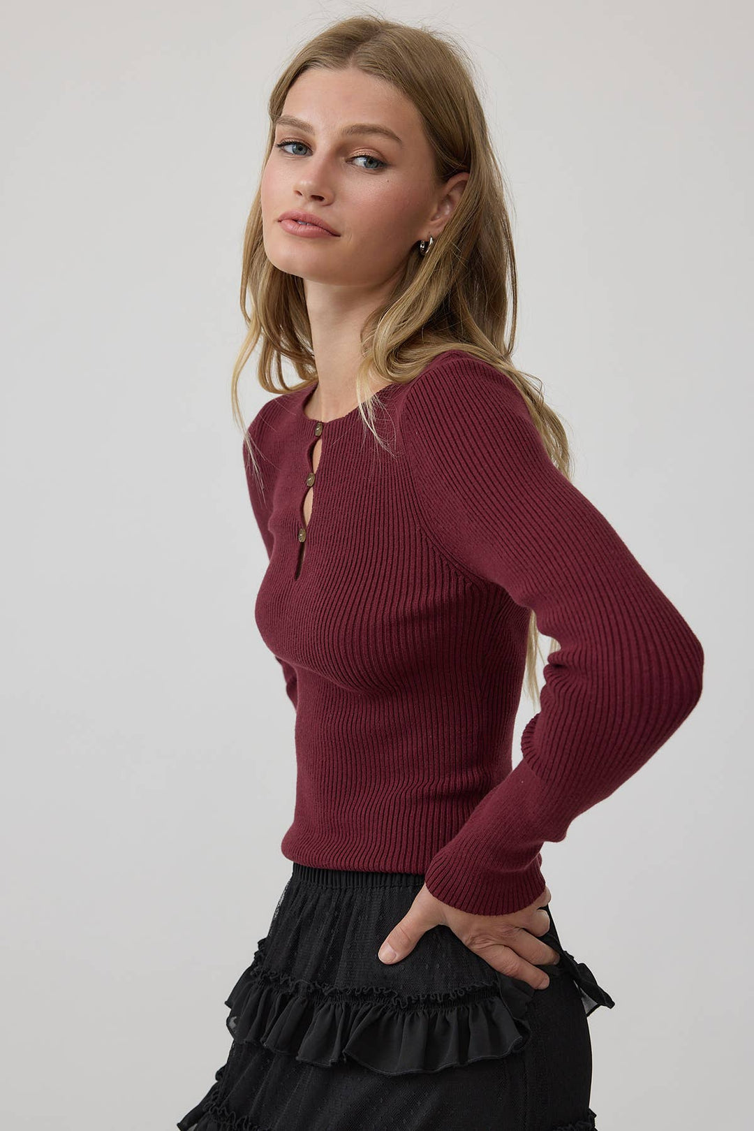 A woman wearing a basic ribbed long-sleeve henley top in deep burgundy, paired with a black ruffled skirt, photographed against a plain light gray studio background.