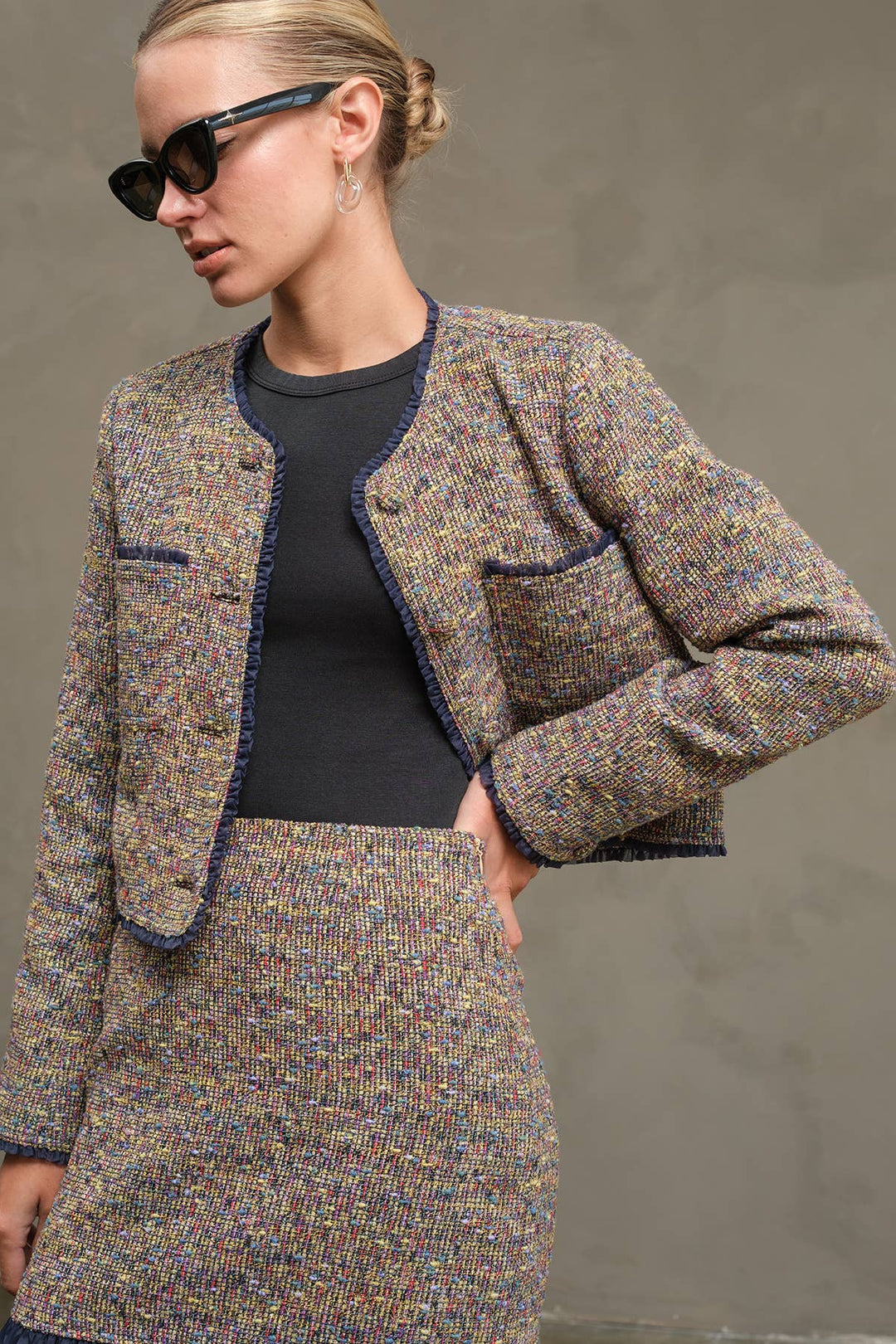 A woman wearing a tweed jacket and matching skirt set in a multicolor blend with dark blue trim, styled over a black top and accessorized with black sunglasses.