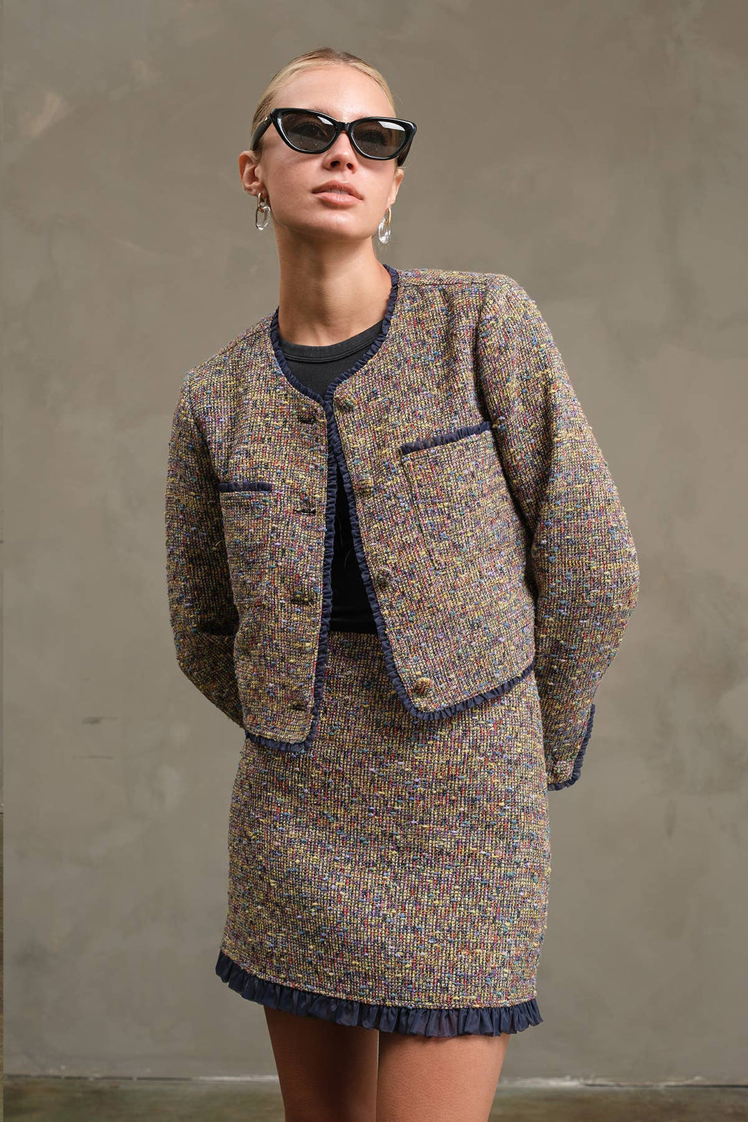 A model wearing a tweed mini skirt and cropped jacket set in a brown, beige, and blue multicolor weave, styled with a dark navy top, sunglasses, and hoop earrings against a textured gray wall.