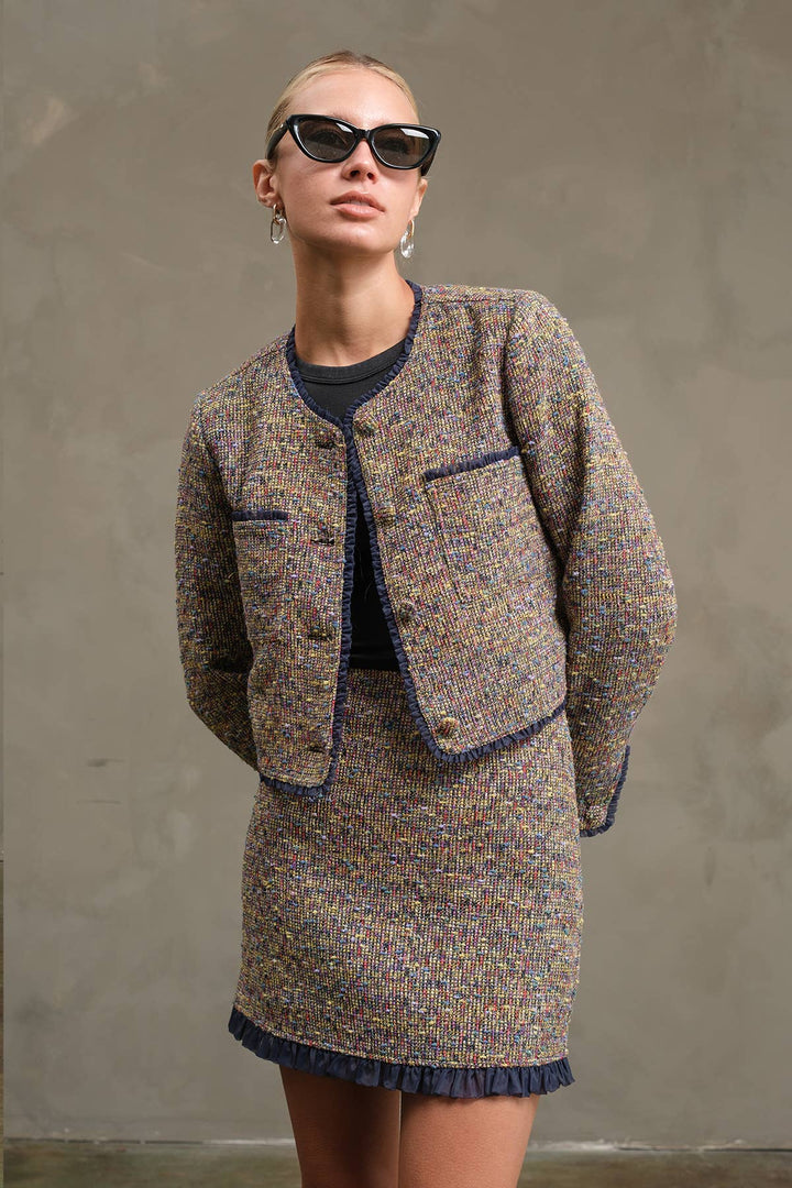 A model wearing a tweed mini skirt and cropped jacket set in a brown, beige, and blue multicolor weave, styled with a dark navy top, sunglasses, and hoop earrings against a textured gray wall.