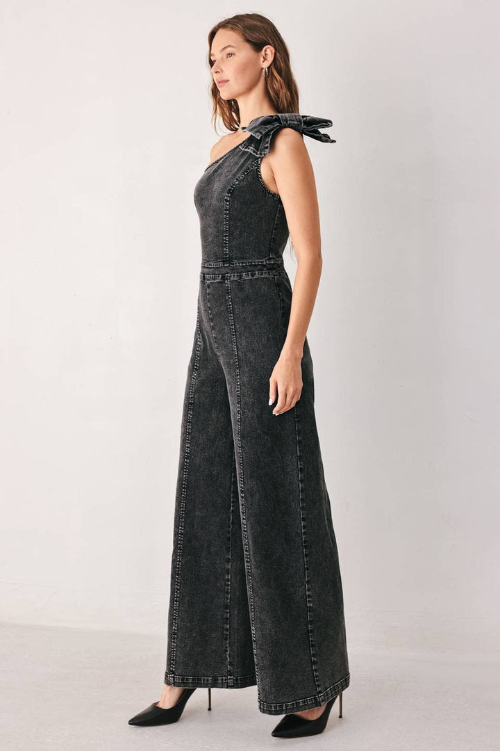 A woman modeling a dark gray washed denim one-shoulder jumpsuit with a bow shoulder detail and wide legs, paired with black pointed-toe heels, against a light gray studio background.