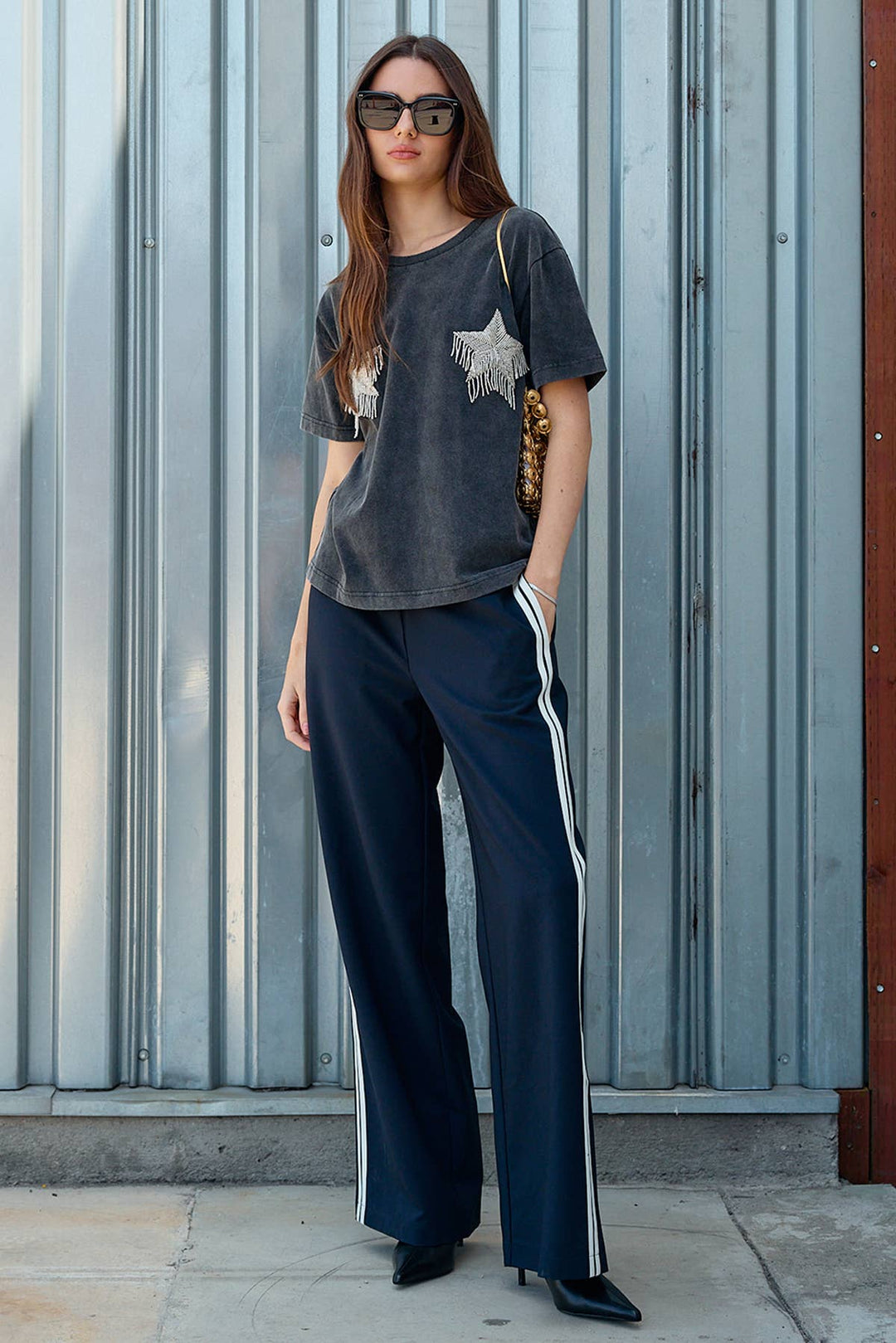 Woman wearing a dark t-shirt with a star design and navy pants with white stripes, standing against a metallic wall.