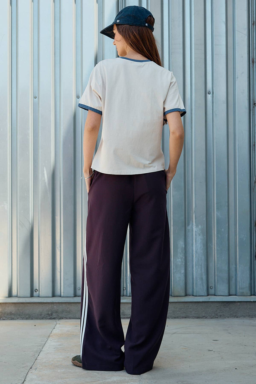 A woman is shown from behind wearing a cream ringer t-shirt with blue trim, dark purple wide-leg sweatpants with white side stripes, and a dark blue baseball cap, standing in front of a corrugated metal wall.