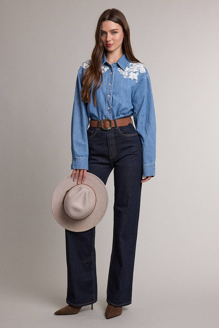 A woman modeling a light blue denim western-style shirt with white floral embroidery on the shoulders, paired with dark blue straight-leg jeans, a brown belt, and holding a beige wide-brim hat against a solid light gray studio background.