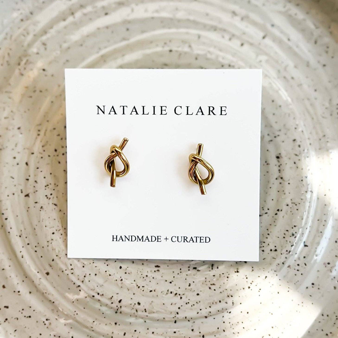 Gold knot earrings on a 'Natalie Clare' branded card against a speckled background