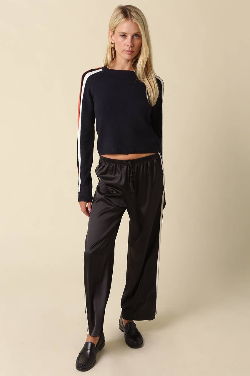 A blonde model wearing a navy long-sleeve crop top with white and red stripes on the sleeves, paired with black satin wide-leg pants and black loafers, against a solid beige background.