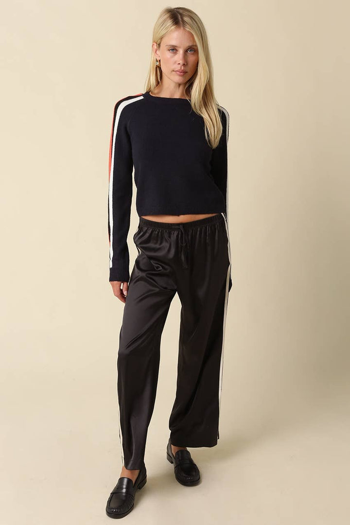 A blonde model wearing a navy long-sleeve crop top with white and red stripes on the sleeves, paired with black satin wide-leg pants and black loafers, against a solid beige background.