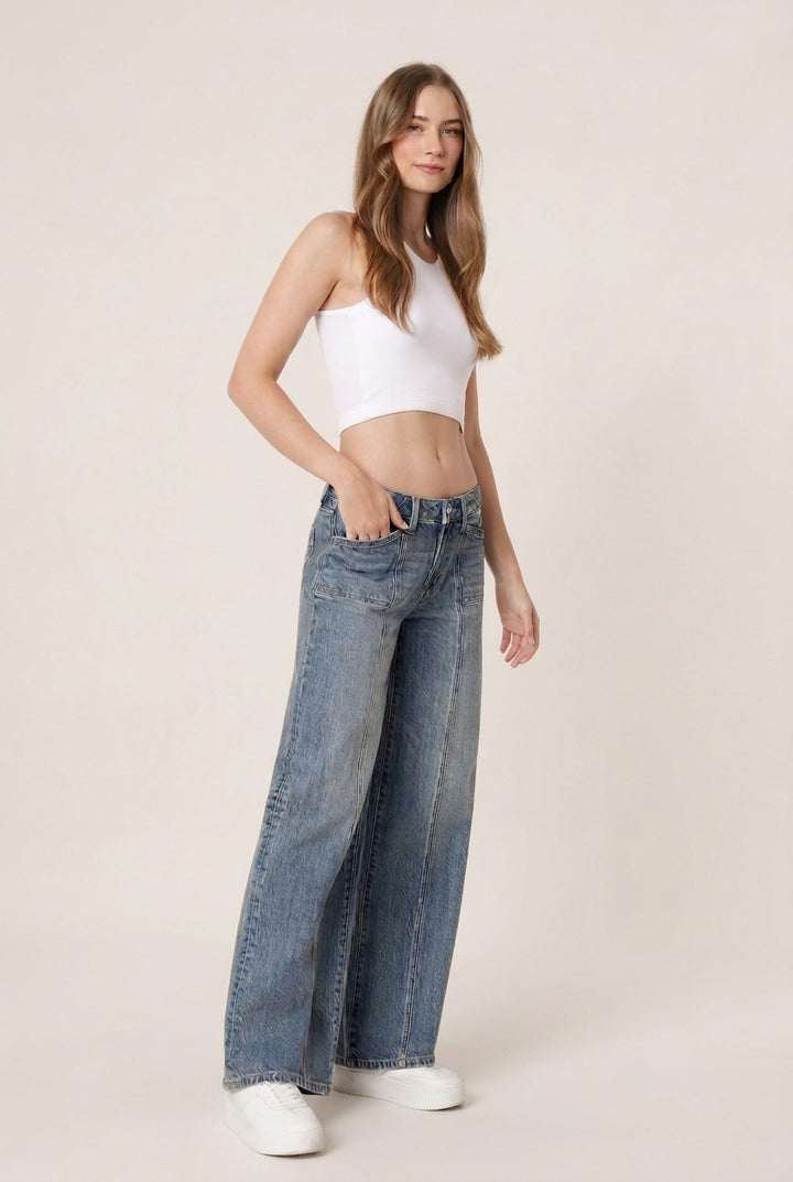 A woman modeling a pair of wide-leg blue jeans with a white crop top and white sneakers, standing against a plain white studio background.