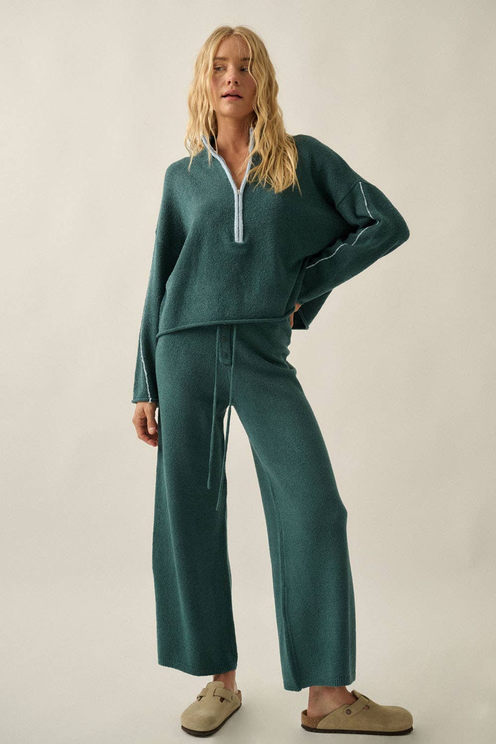 A woman modeling a matching two-piece lounge set in a deep teal green color, featuring a long-sleeve half-zip pullover and wide-leg pants, styled with beige slip-on shoes against a plain light beige studio background.