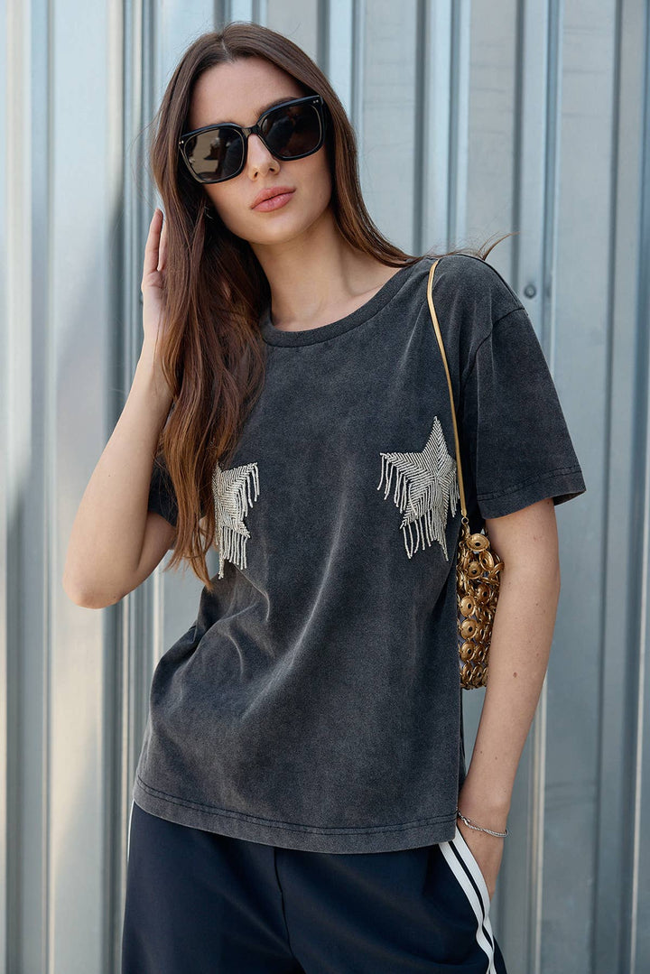Woman wearing a dark gray t-shirt with fringe details against a metallic wall.
