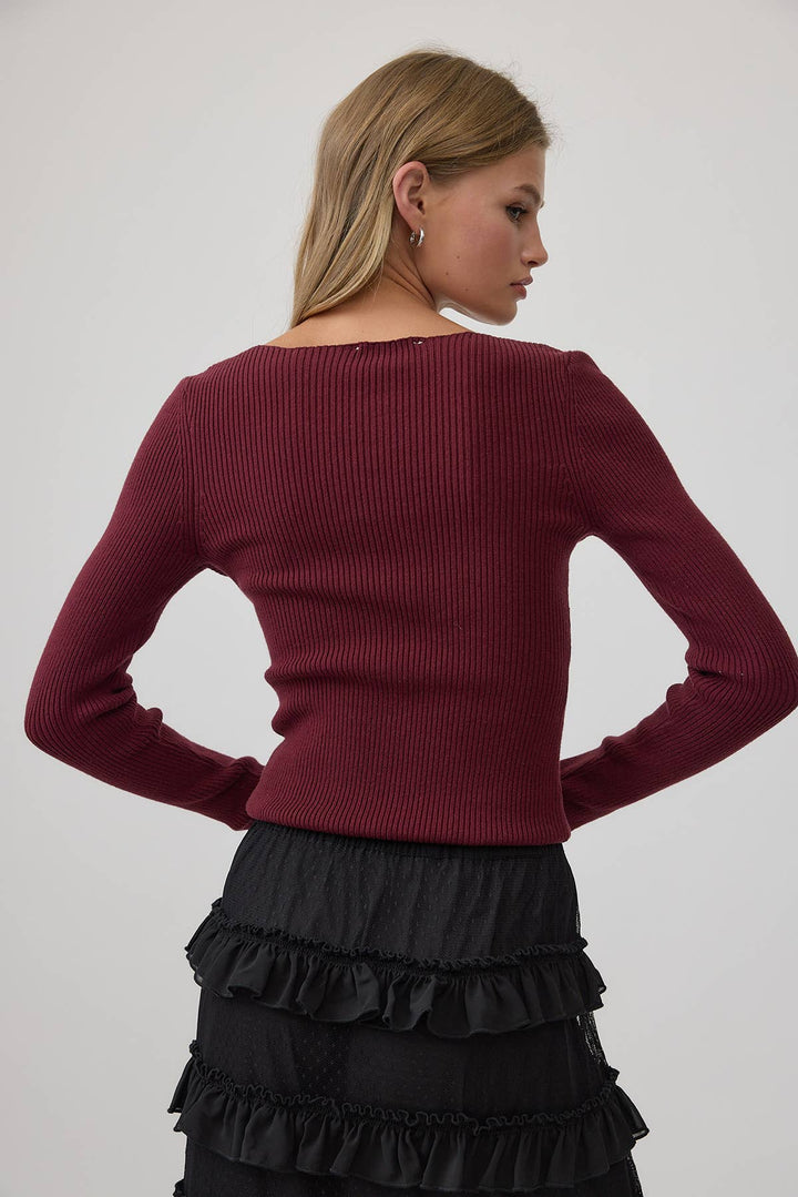 A woman shown from the back wearing a ribbed long-sleeve top in deep burgundy and a black ruffled skirt, against a plain light gray studio background.
