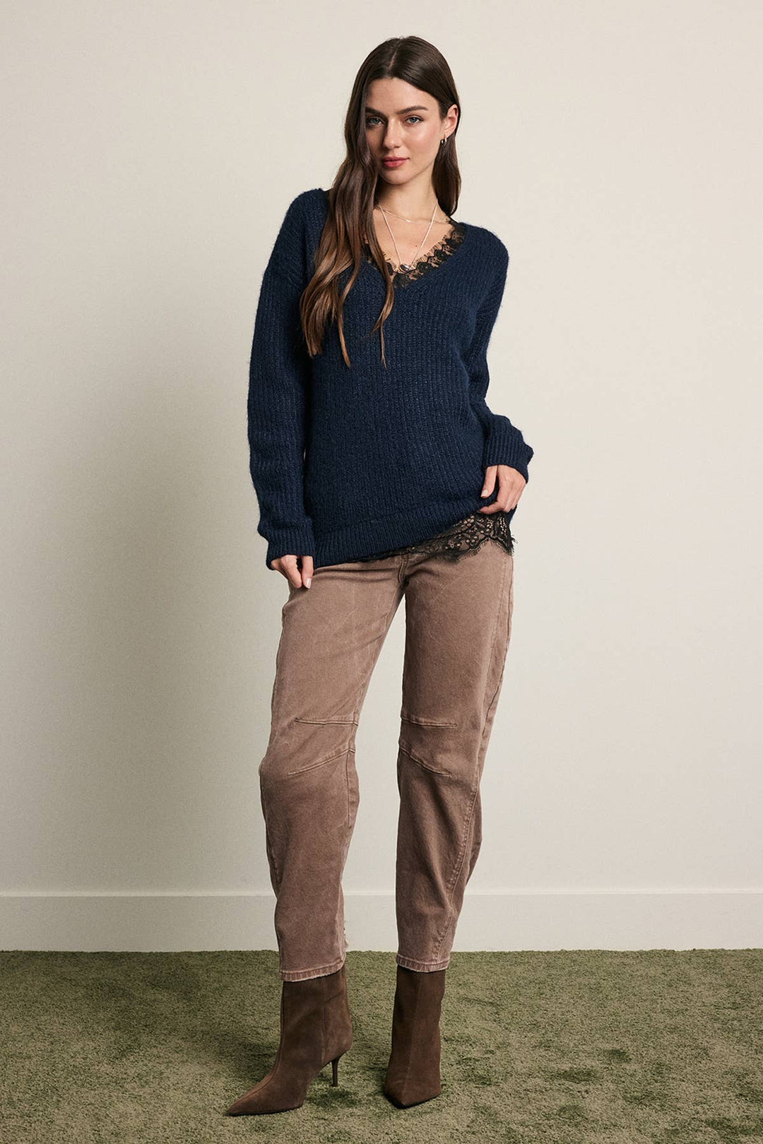 A woman modeling a navy blue knit sweater with a lace-trimmed neckline, paired with brown pants and brown ankle boots, standing against a plain beige wall.
