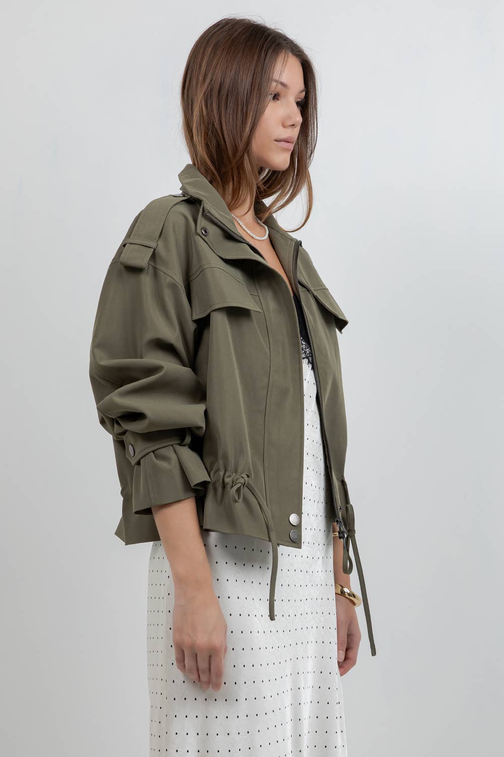 Woman wearing an olive green jacket over a white dress on a plain background