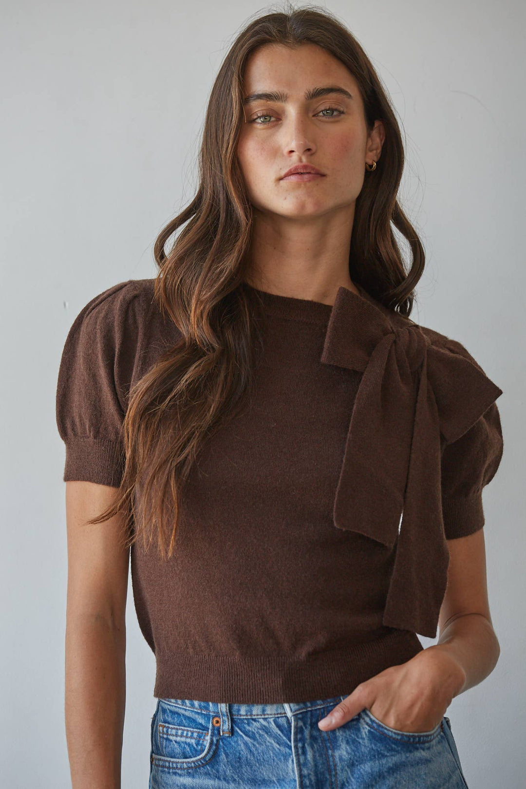 A woman wearing a brown short-sleeve knit top with puff sleeves, a crew neckline, and a large bow detail on the right side of the chest, paired with blue jeans against a plain light gray background.