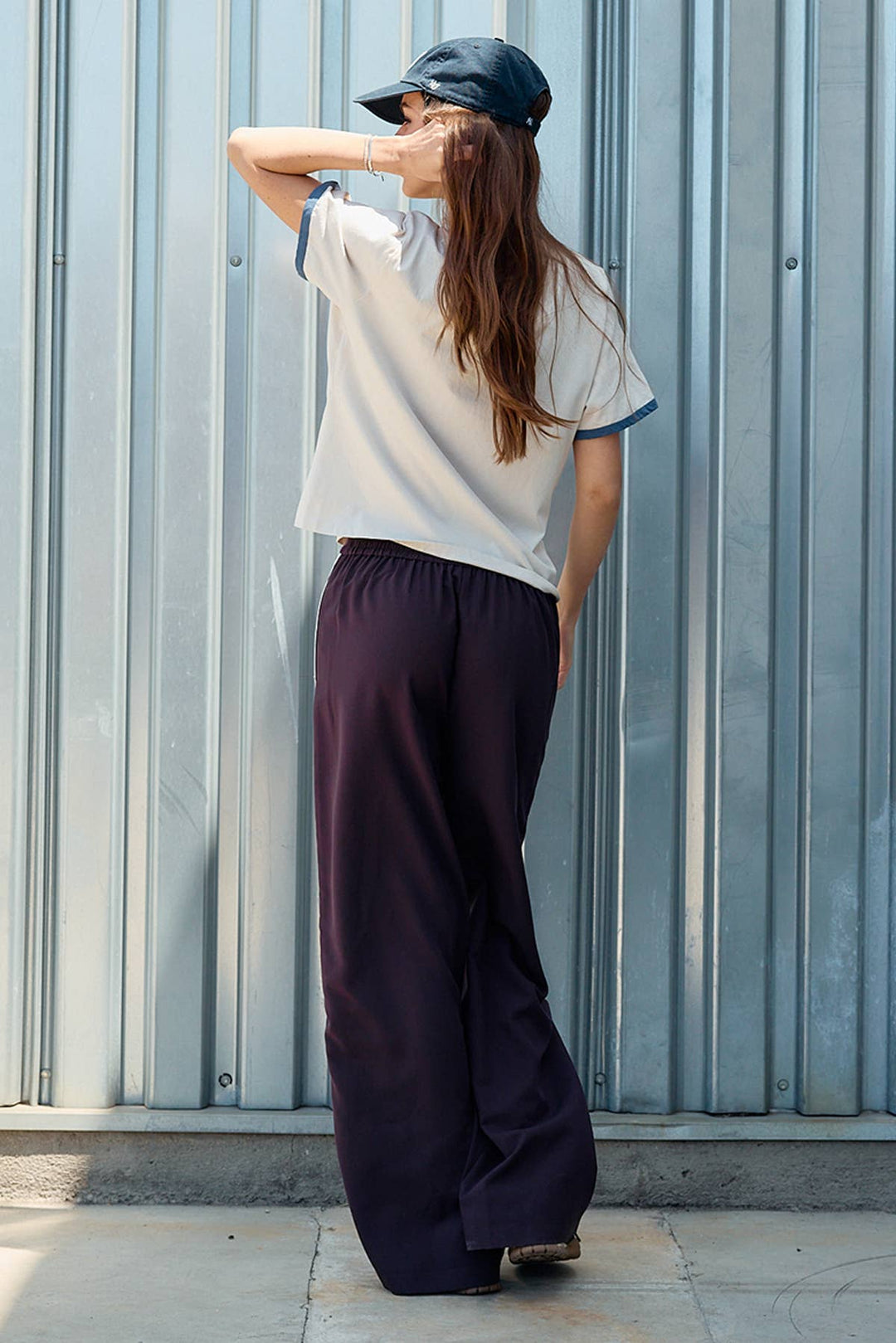 A woman stands with her back to the camera in front of a corrugated metal wall, wearing a white ringer T-shirt with blue trim, maroon colored wide-leg pants, and a dark cap.
