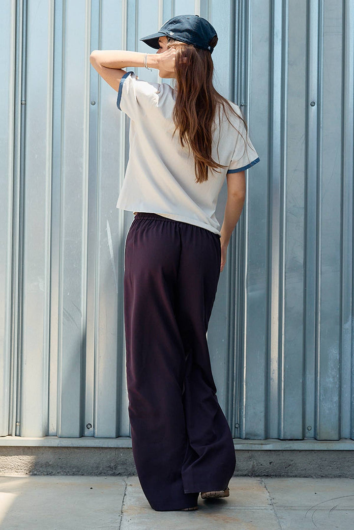 A woman stands with her back to the camera in front of a corrugated metal wall, wearing a white ringer T-shirt with blue trim, maroon colored wide-leg pants, and a dark cap.