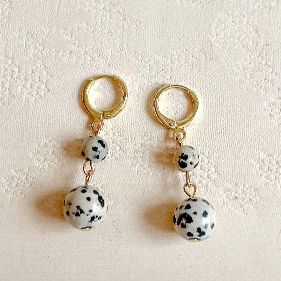 A pair of gold hoop dangle earrings with white beads speckled like Dalmatian spots, displayed on a textured cream fabric background.