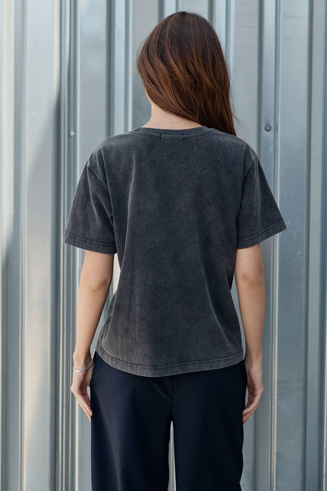 Person wearing a dark gray t-shirt and black pants standing against a corrugated metal wall.