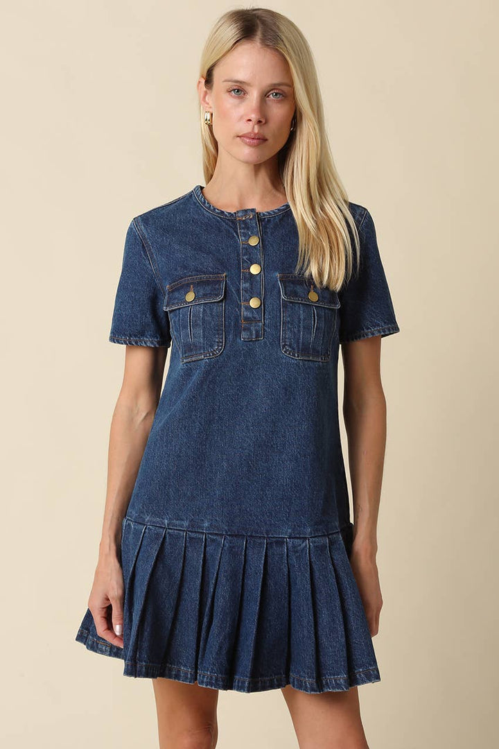 A blonde model wearing a short-sleeve denim dress with a buttoned henley neckline and pleated skirt, set against a solid beige background.